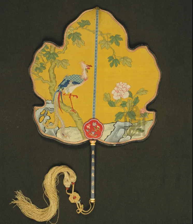 Yellow leaf-shaped Kesi silk fan featuring a colorful phoenix perched on a Wutong branch next to blooming peonies and garden rocks.