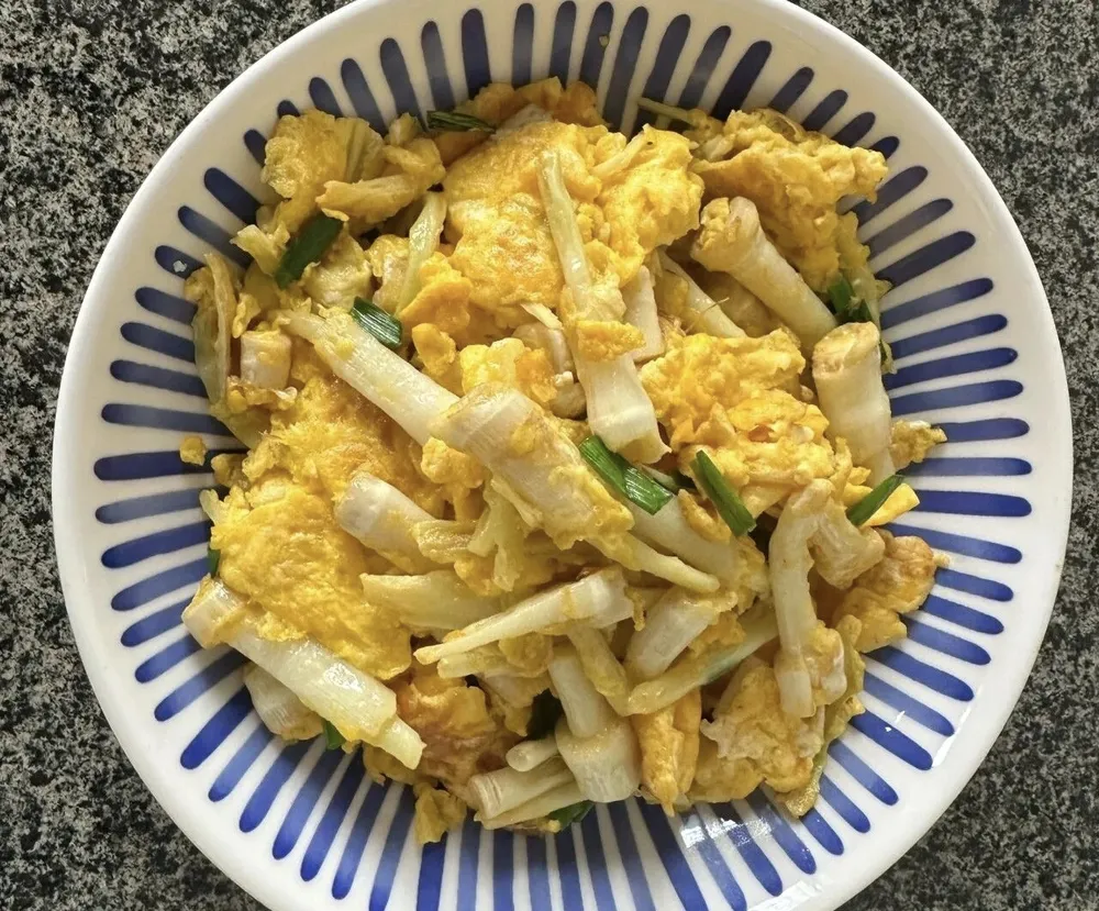 A home-cooked dish of stir-fried tender white cattail shoots (pucai) and golden scrambled eggs served in a blue-striped ceramic bowl.